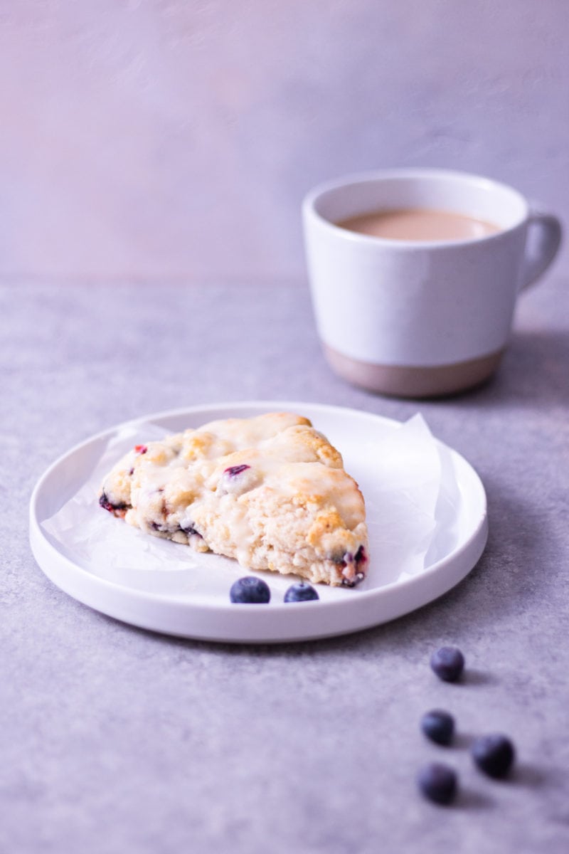Angled view of a lemon blueberry scone on a plate surrounded by scattered fresh blueberries with a mug of coffee in the background on a light grey surface.