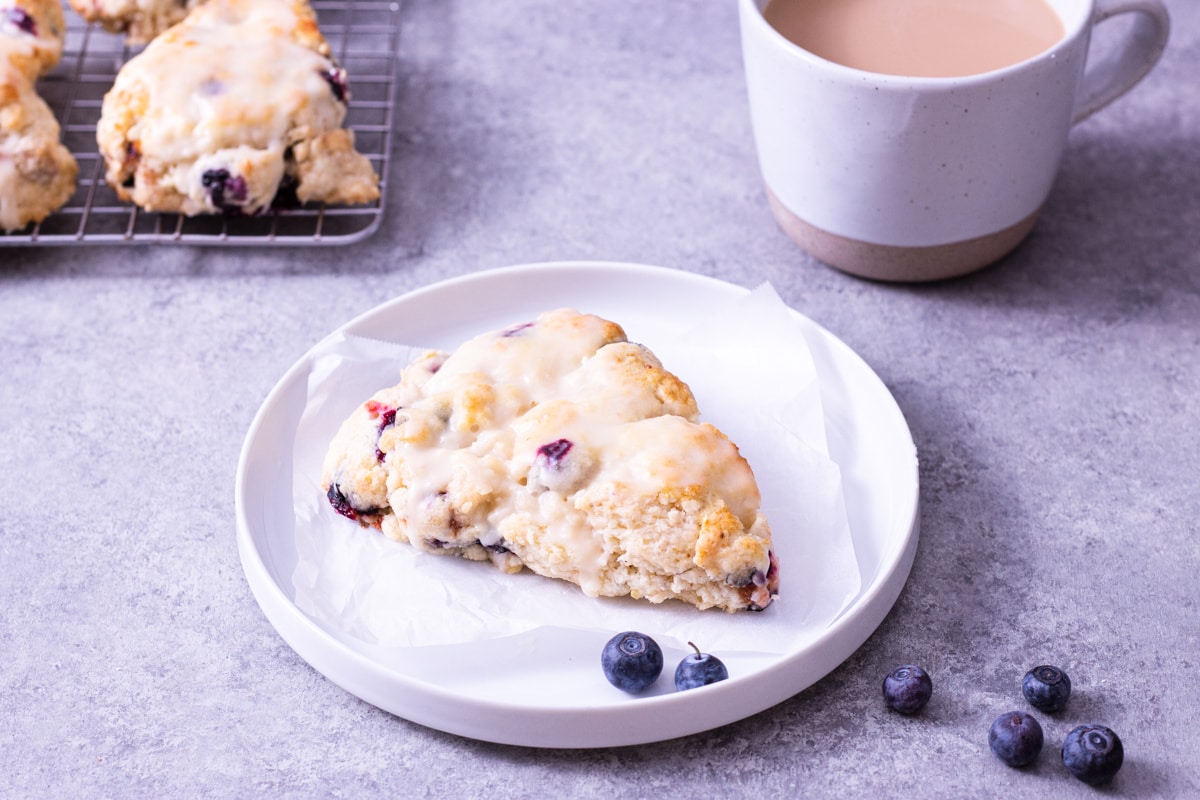 Angled view of a lemon blueberry scone on a plate surrounded by scattered fresh blueberries with a mug of coffee and more scones in the background on a light grey surface.