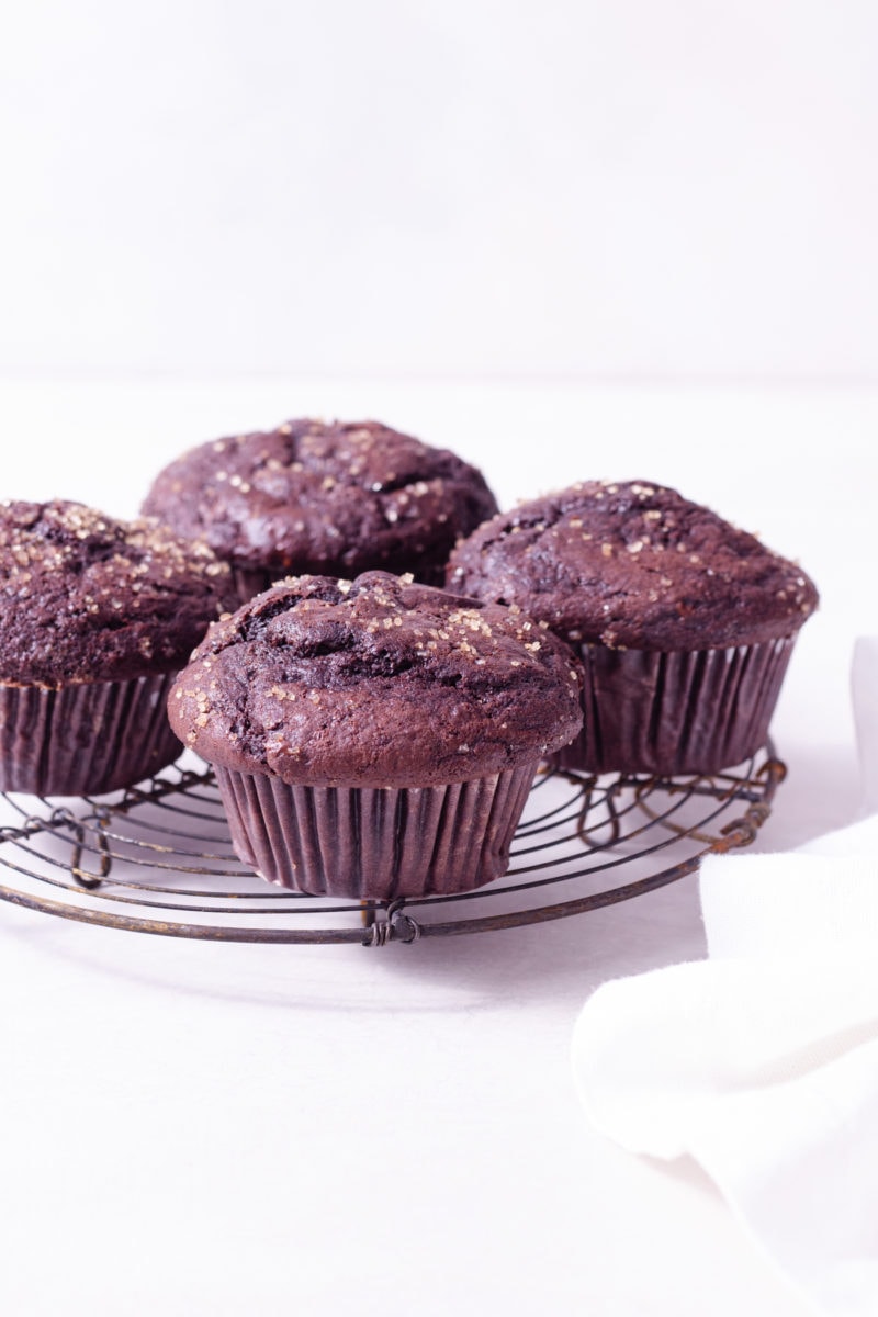 Straight on shot of Double Chocolate Chip Muffins on a wire rack next to a dish cloth on a white surface.