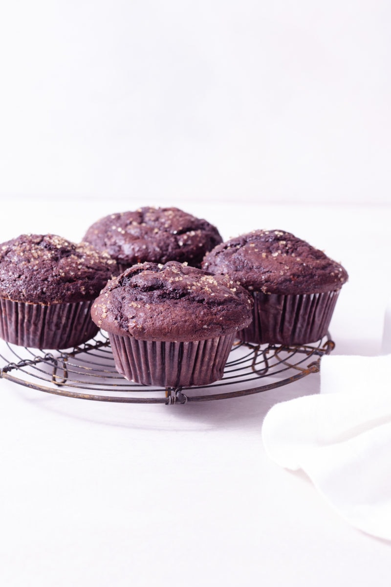 Straight on shot of Double Chocolate Chip Muffins on a wire rack next to a dish cloth on a white surface.