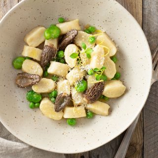 Overhead view of a bowl of Homemade Gnocchi with Morel Mushrooms, Peas and Fava Beans on a light wood surface.