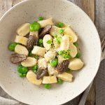 Overhead shot of a bowl of gnocchi with spring vegetables on a light, wood surface.