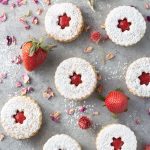 Overhead shot of linzer cookies dusted with powdered sugar and filled with strawberry rose jam, surrounded by fresh strawberries and dried rose petals on a light grey textured surface.