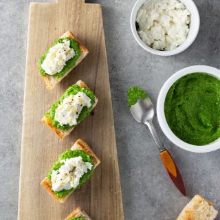 Overhead shot of crostini topped with ramp pesto and ricotta cheese on a light wood cutting board, surrounded by bowls of ricotta and ramp pesto, a spoon and slices of toast on a grey, textured surface.