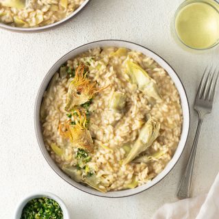 Overhead shot of 2 bowls of artichoke risotto garnished with gremolata and fried artichoke slices, surrounded by 2 glasses of white wine, a fork and a dinner napkin.
