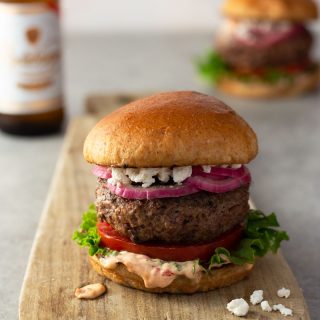 Straight on shot of a harissa lamb burger with spicy mayonnaise, lettuce, tomato, quick pickled red onions and feta on a brioche bun on a light wood cutting board on a light grey surface with a beer and burger in the background.