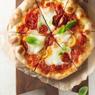 Overhead shot of a margherita pizza topped with colored, heirloom tomatoes, pizza sauce and buffalo mozzarella cheese on parchment on a dark wood French cutting board on an off white, textured surface.