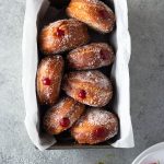 Overhead shot of homemade jelly doughnuts with strawberry jam in a loaf pan surrounded by dessert plates and fresh strawberries on a light grey, textured surface.