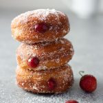 Straight on shot of a stack of homemade jelly doughnuts with strawberry jam, coated in sugar on a light grey surface surrounded by fresh strawberries with a coffee cup in the background.