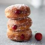Straight on shot of a stack of homemade strawberry jam doughnuts, coated in sugar on a light grey surface surrounded by fresh strawberries with a coffee cup in the background.