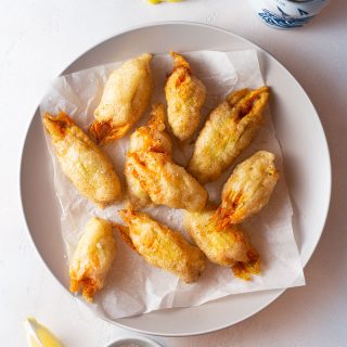 Overhead shot of a plate of fried squash blossoms stuffed with crab and mascarpone cheese on a light textured surface, surrounded by lemon wedges, a dish of salt and a beer.