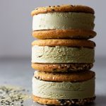 Straight on shot of a stack of matcha green tea ice cream sandwiches with sesame tahini cookies on a light grey surface surrounded by black and white sesame seeds and matcha powder.