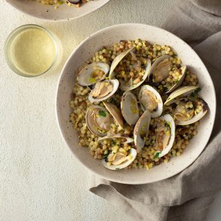 Overhead shot of two light, rustic bowls of fregola pasta with clams surrounded by a glass of white wine and a neutral colored towel on a light, cream colored, textured plaster surface.