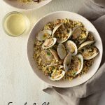 Overhead shot of two light, rustic bowls of fregola pasta with clams surrounded by a glass of white wine and a neutral colored towel on a light, cream colored, textured plaster surface.