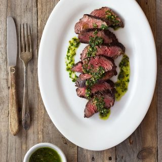 Overhead shot of sliced hanger steak drizzled in an Italian salsa verde on a light, rustic wood surface with rustic utensils and a small bowl of salsa verde.