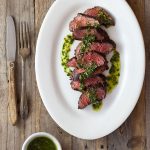 Overhead shot of sliced hanger steak drizzled in an Italian salsa verde on a light, rustic wood surface with rustic utensils and a small bowl of salsa verde.