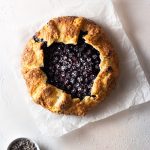 Overhead shot of a lavender blueberry galette on parchment paper on a light, textured background next to a small bowl of dried lavender.
