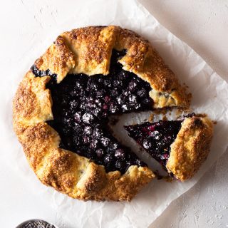 Overhead shot of a lavender blueberry galette with a slice cut out on parchment paper on a light, textured background next to a small bowl of dried lavender.