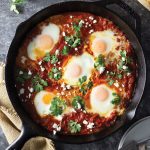 Overhead shot of a cast iron pan of shakshuka (baked eggs in a spiced tomato sauce) with harissa paste, feta cheese and cilantro on a dark grey textured surface surrounded by a dish towel, white plates and forks, and sliced crusty bread.