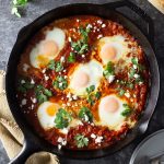Overhead shot of a cast iron pan of shakshuka (baked eggs in a spiced tomato sauce) with harissa paste, feta cheese and cilantro on a dark grey textured surface surrounded by a dish towel and sliced crusty bread.