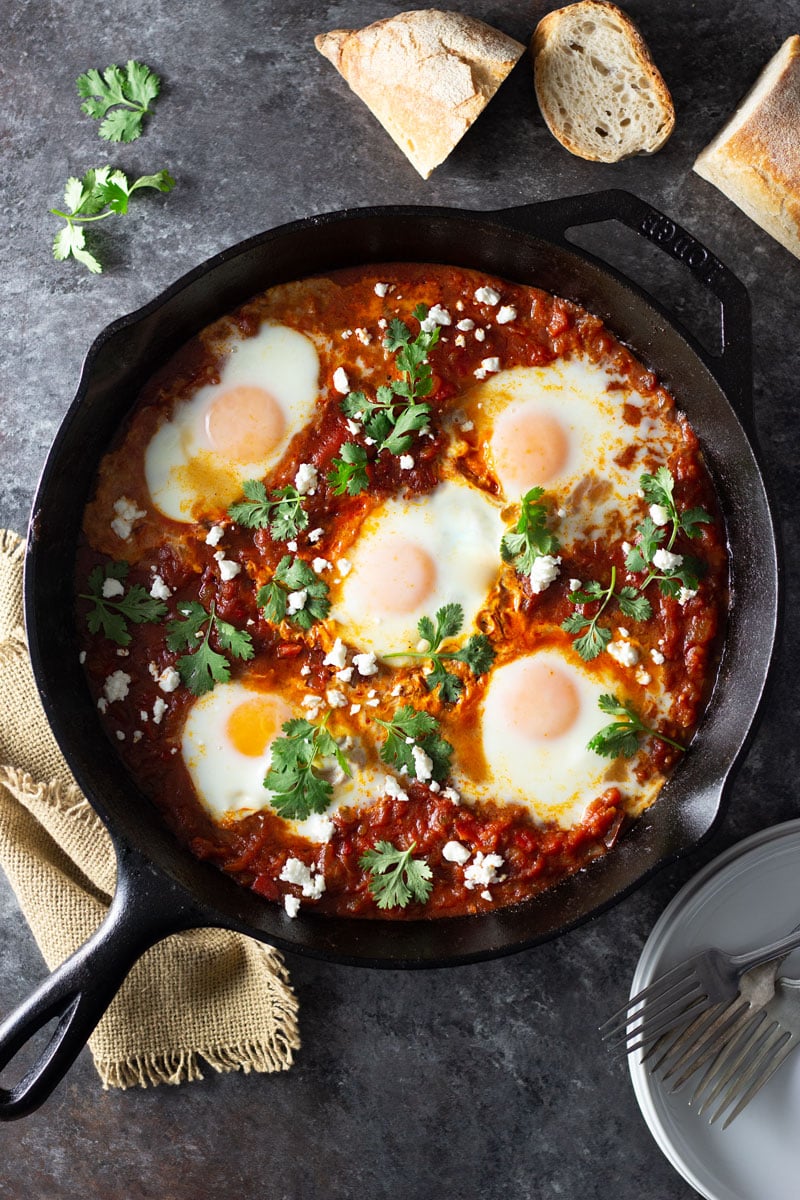 Overhead shot of a cast iron pan of shakshuka (baked eggs in a spiced tomato sauce) with harissa paste, feta cheese and cilantro on a dark grey textured surface surrounded by a dish towel, white plates and forks, and sliced crusty bread.
