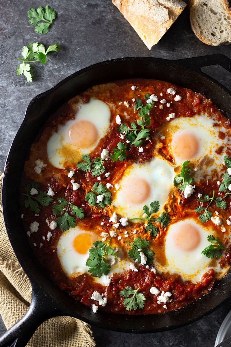 Overhead shot of a cast iron pan of shakshuka (baked eggs in a spiced tomato sauce) with harissa paste, feta cheese and cilantro on a dark grey textured surface surrounded by a dish towel, and sliced crusty bread.