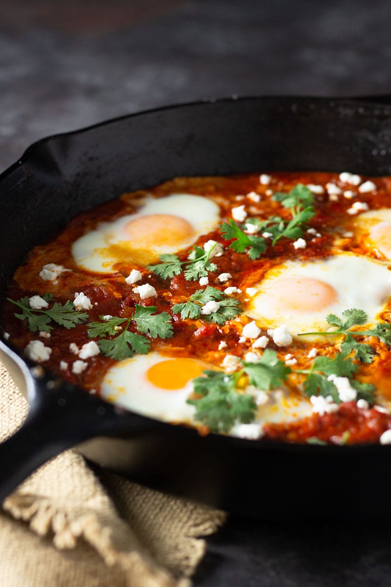 ¾ angled shot of a cast iron pan of shakshuka (baked eggs in a spiced tomato sauce) with harissa paste, feta cheese and cilantro on a dark grey textured surface next to a tan dish towel.