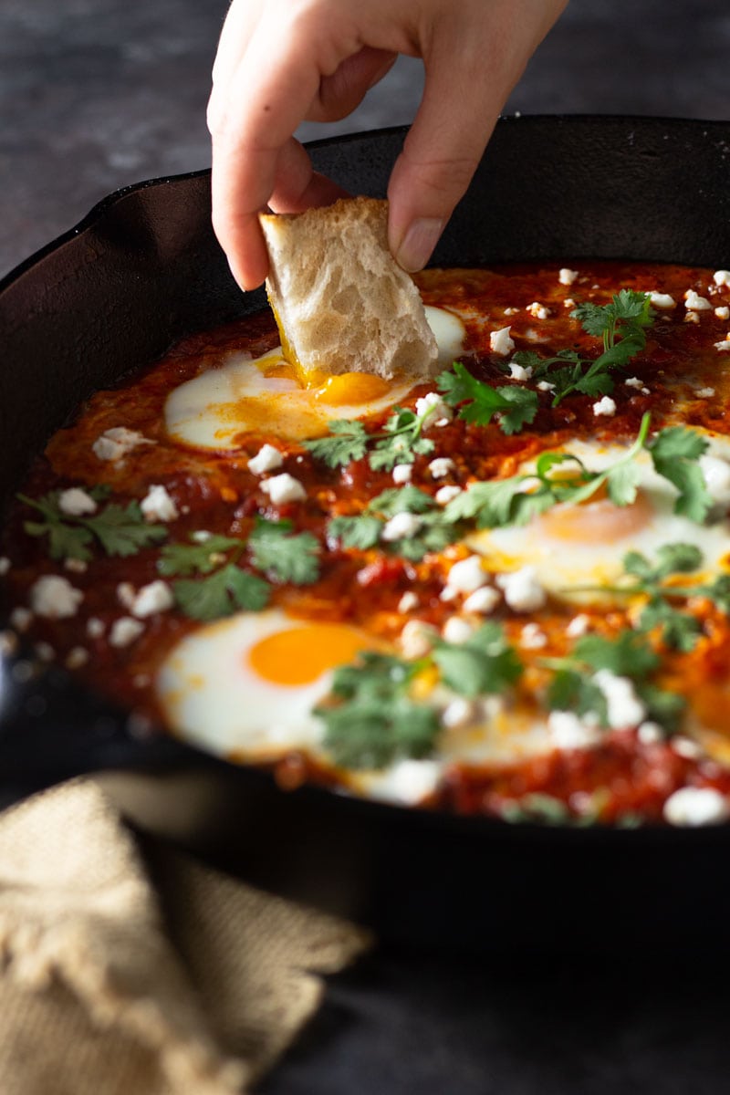 ¾ angled shot of a cast iron pan of shakshuka (baked eggs in a spiced tomato sauce) with harissa paste, feta cheese and cilantro with a hand dipping a piece of bread into the yolk on a dark grey textured surface next to a tan dish towel.