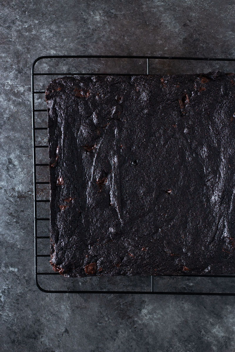 Overhead shot of dark chocolate chunk brownies on a cooling rack on a dark, rustic textured background.