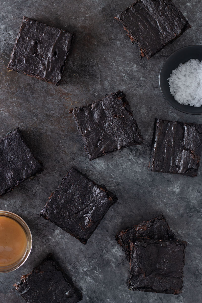 Overhead shot of dark chocolate chunk brownies surrounded by a small bowl of salt and a small bowl of caramel on a dark, rustic textured background.