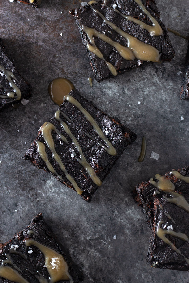 Overhead shot of dark chocolate chunk brownies drizzled with tamarind salted caramel and topped with maldon salt on a dark, rustic textured background.
