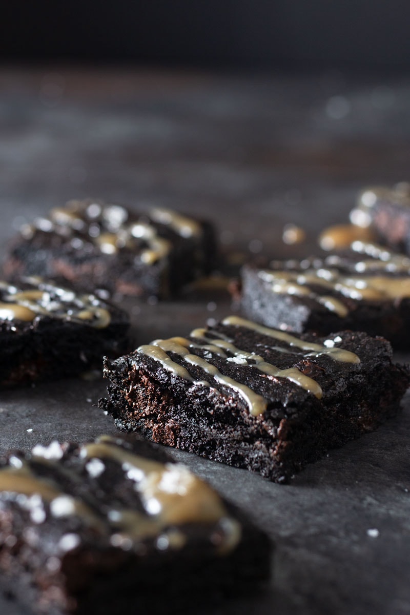 Straight on shot of dark chocolate chunk brownies drizzled with tamarind salted caramel and topped with maldon salt on a dark, rustic textured background.