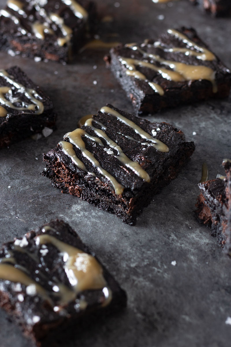 ¾ angled shot of dark chocolate chunk brownies drizzled with tamarind salted caramel and topped with maldon salt on a dark, rustic textured background.