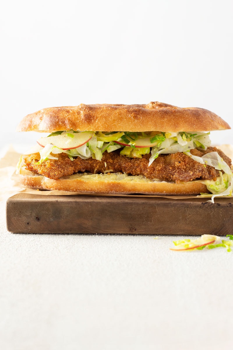 Straight on shot of a Pork Schnitzel Sandwich with Apple Brussels Sprout Slaw on a mini focaccia bread on parchment paper on a wooden cutting board on a white plaster surface with a white background.