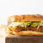 Straight on shot of a Pork Schnitzel Sandwich with Apple Brussels Sprout Slaw on a mini focaccia bread on parchment paper on a wooden cutting on a white plaster surface with a white background.