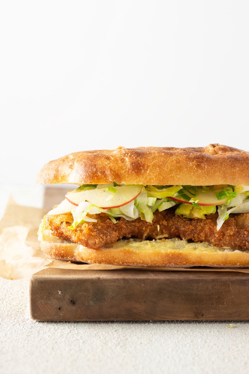 Straight on shot of a Pork Schnitzel Sandwich with Apple Brussels Sprout Slaw on a mini focaccia bread on parchment paper on a wooden cutting on a white plaster surface with a white background.