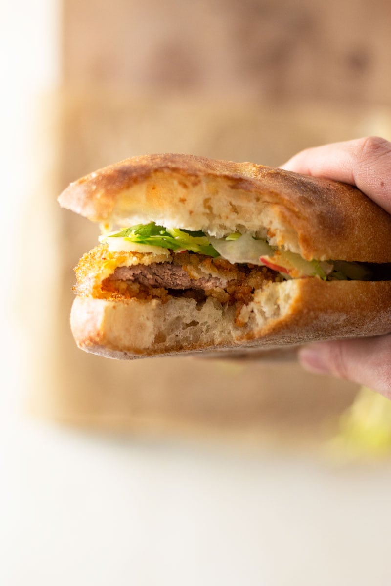 Close up shot of a hand holding a Pork Schnitzel Sandwich with Apple Brussels Sprout Slaw and homemade dijonnaise with a bite taken out of it above a wooden cutting board and white surface. 
