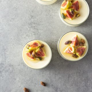 Overhead shot of four glasses of buttermilk goat cheese panna cotta topped with figs, pistachios and honey on a light, grey textured surface.