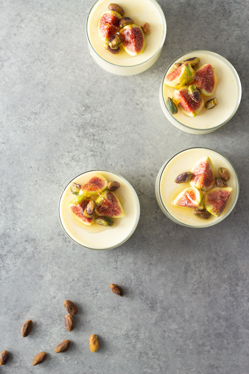 Overhead shot of four glasses of buttermilk goat cheese panna cotta topped with figs, pistachios and honey on a light, grey textured surface.