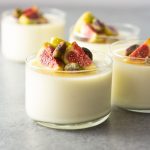 Straight on shot of four glasses of buttermilk goat cheese panna cotta topped with figs, pistachios and honey on a light, grey textured surface.