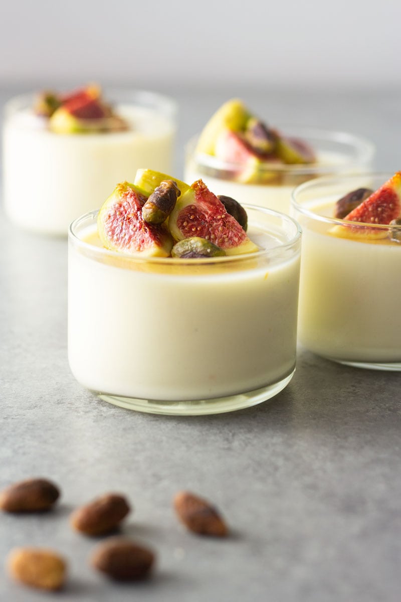 Straight on shot of four glasses of buttermilk goat cheese panna cotta topped with figs, pistachios and honey on a light, grey textured surface.
