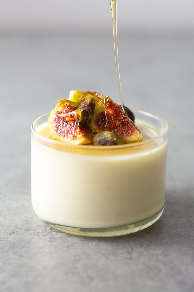 Straight on shot of honey drizzling over a glass of buttermilk goat cheese panna cotta topped with figs and pistachios on a light, grey textured surface.