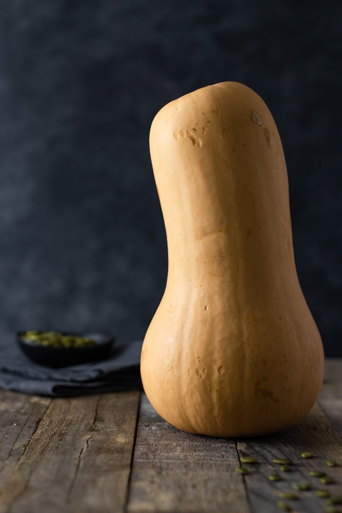 Straight on shot of a whole raw butternut squash standing up on a grey wood surface with a dark textured background with a small bowl of pepitas and a dish towel in the background.