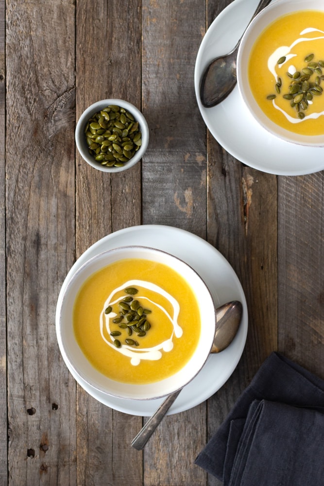 Overhead view of two bowls of creamy butternut squash soup topped with cream and pepitas in white bowls over a white plate with rustic spoons surrounded by a small bowl of pepitas and a dark dish towel on a rustic grey wood surface.
