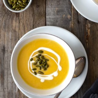 Overhead view of two bowls of creamy butternut squash soup topped with cream and pepitas in white bowls over a white plate with rustic spoons surrounded by a small bowl of pepitas and a dark dish towel on a rustic grey wood surface.
