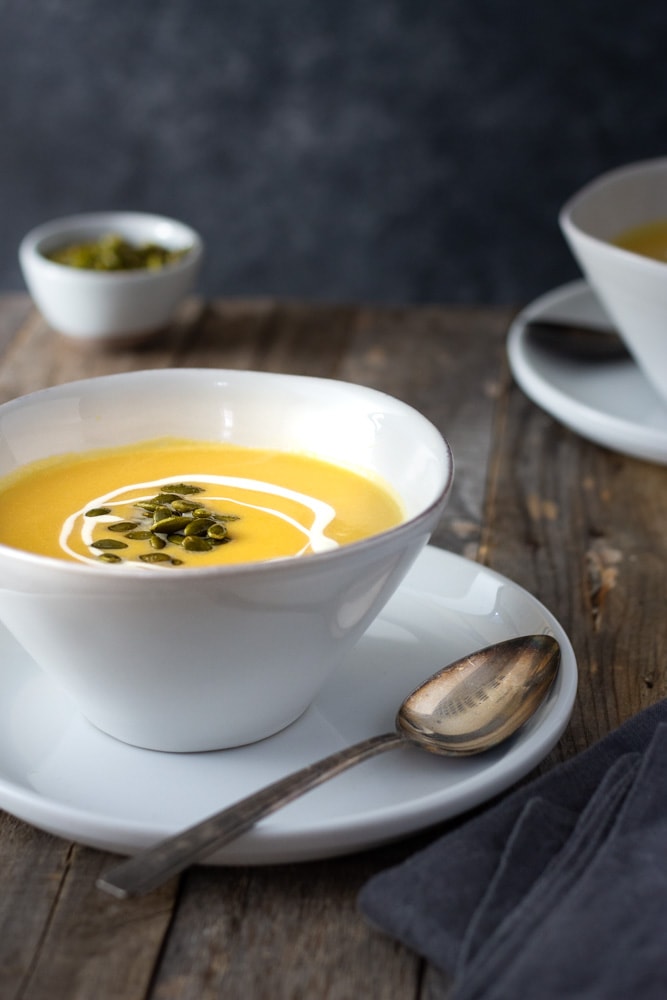 ¾ angled view of two bowls of creamy butternut squash soup topped with cream and pepitas in white bowls over a white plate with rustic spoons surrounded by a small bowl of pepitas and a dark dish towel on a rustic grey wood surface with a dark textured background.
