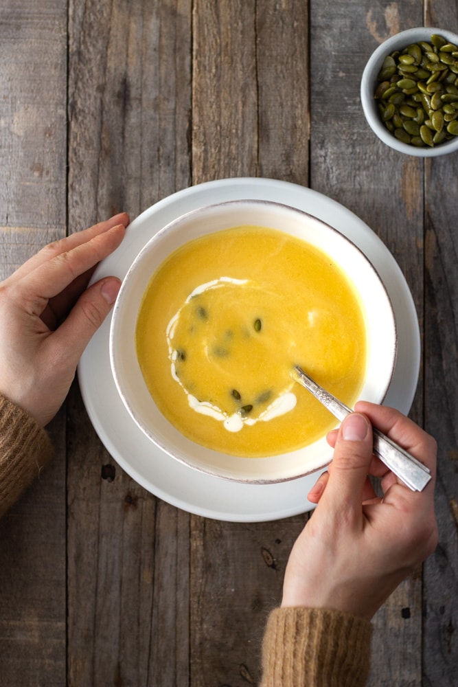 Overhead view of hands ready to eat a bowl of creamy butternut squash soup in a white bowl over a white plate next to a small bowl of pepitas on a grey rustic wood surface.
