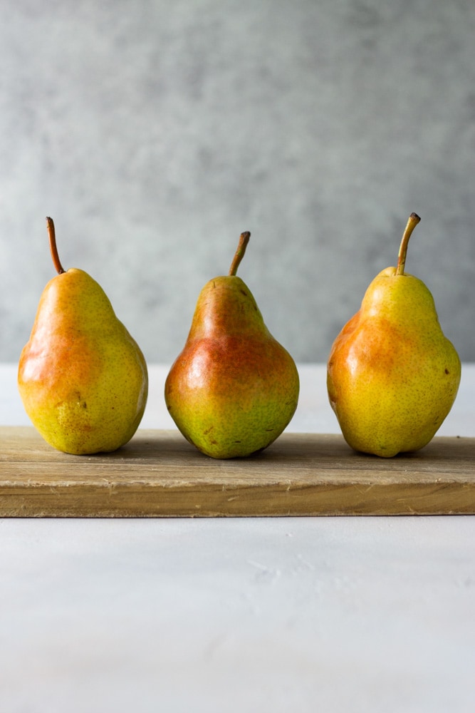 Straight on view of three upright bartlett pears on a light wood cutting board on an off white textured surface with a light grey textured background.