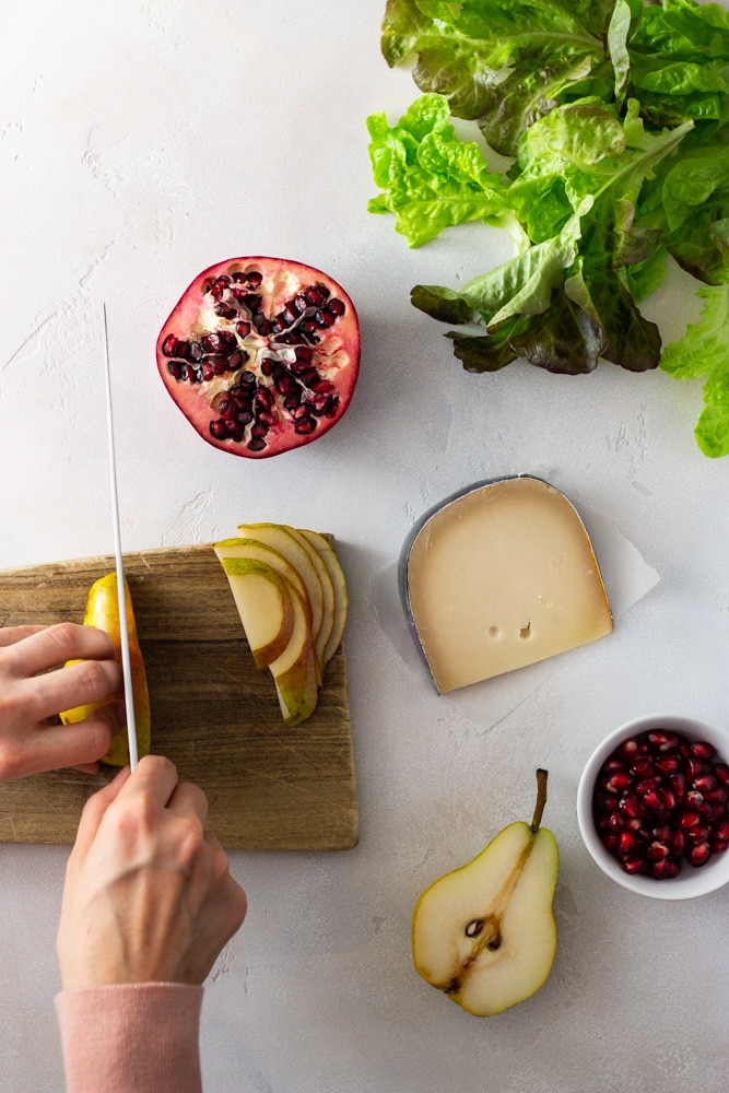Overhead view of hands cutting a pear on a light wooden cutting board surrounded by a halved pear, a halved pomegranate, a bowl of pomegranate seeds and green and red leaf lettuce on an off white textured surface.