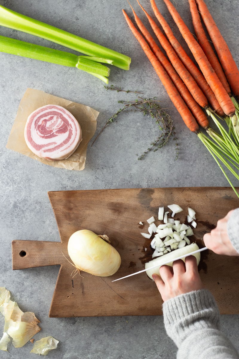 Overhead view of hands chopping an onion on a wooden cutting board, surrounded by pancetta, fresh thyme, celery and carrots on a light grey textured surface. 
